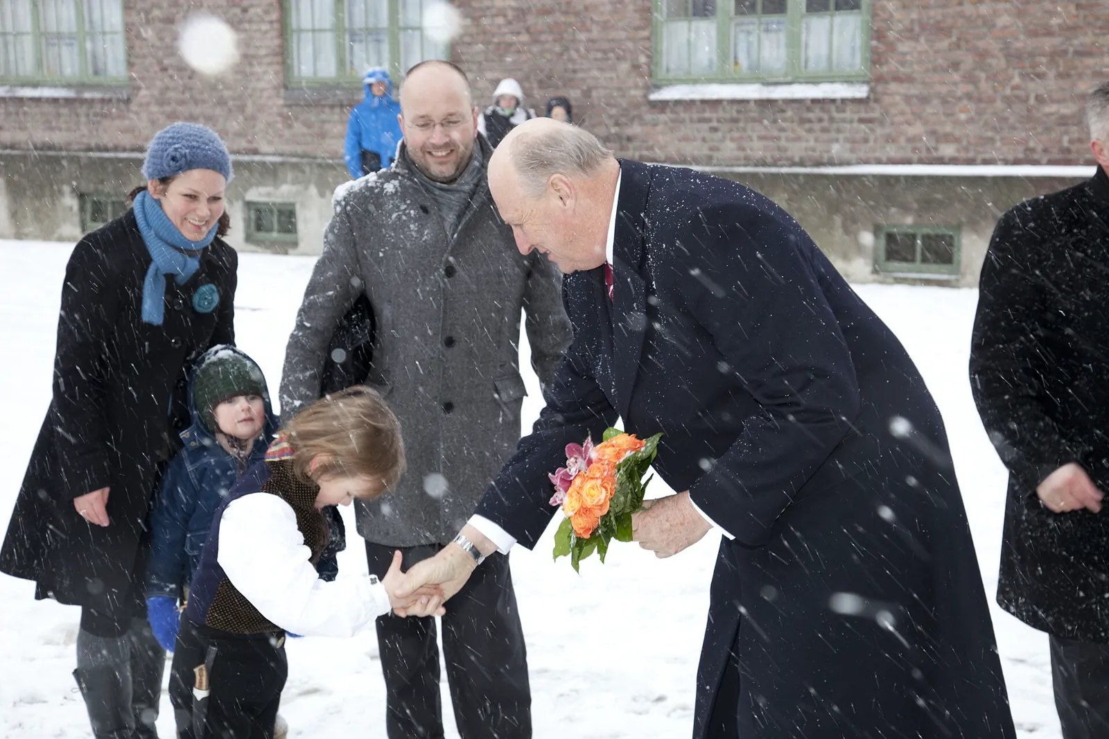 Kong Harald bøyer seg ned i tett snøvær og tar en liten jente i hånden. Han smiler og holder en blomsterbukett.